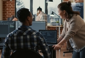 Two people in an office, one is sitting in front of a monitor, the other is leaning on the desk, they are in conversation