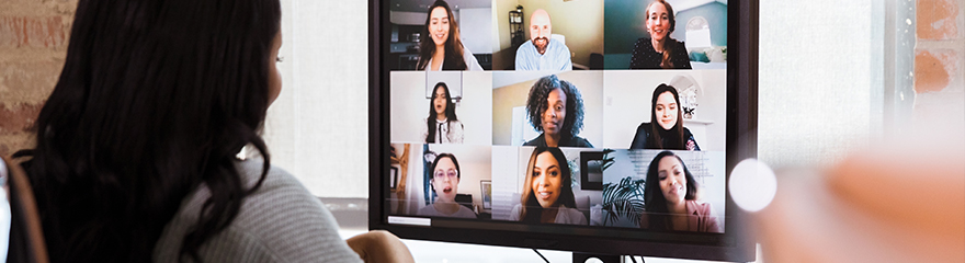 A woman sits at a computer in a virtual meeting. Nine faces appear on her desktop screen.