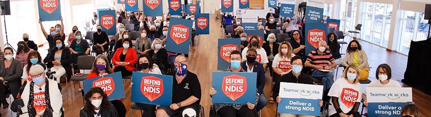 peaceful gathering of people in a hall with signs reading Defend the NDIS