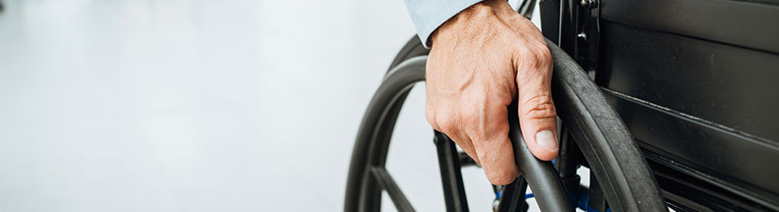 A cropped image of a person in a wheelchair shot from down low, only showing part of the wheel and the person's hand on the wheel