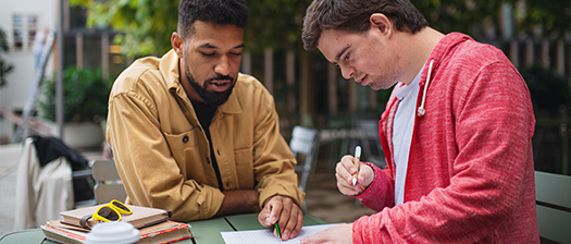 Two people sit at an outdoor table. One is pointing to an piece of paper, the other is writing on it.