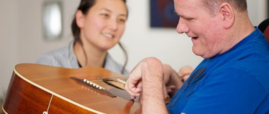 A support worker smiles at a person with disability who is holding a guitar