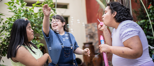 Three young people with disability stand in a courtyard. One is blowing bubbles and the two others are smiling and playing with the bubbles.
