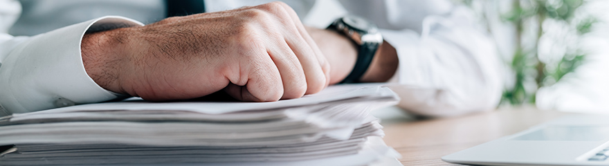 Close up of a fist resting on a pile of papers, the person in the out of focus background is wearing a shirt and tie.