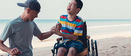 A young person with disability sitting in a wheelchair on a beach having fun with their support person who is standing nearby