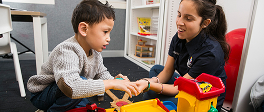 A support worker interacts with a young boy in a room. Lots of toys are in the foreground  