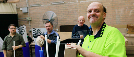 Four men with disability stand in a room. A man in the foreground with a hi-vis vest smiles at the camera
