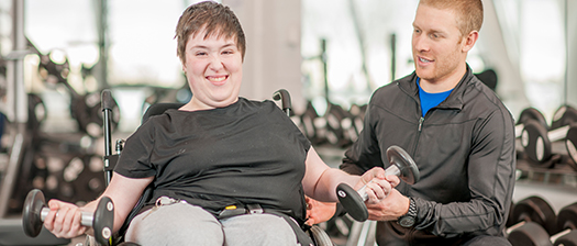 A man helps a woman with disability lift weights in a gym. They both smile.