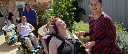 Four women and one man are outside, with three of the women using wheelchairs. They come down a garden path and are all smiling.