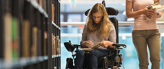 A woman using a wheelchair is in a library reading a book. Another woman is standing next to her, holding a stack of books