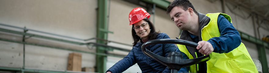 A man with down syndrome wears a hi-vis vest and drives a small vechile around a factory, while a woman wearing a red hard hat sits next to him.