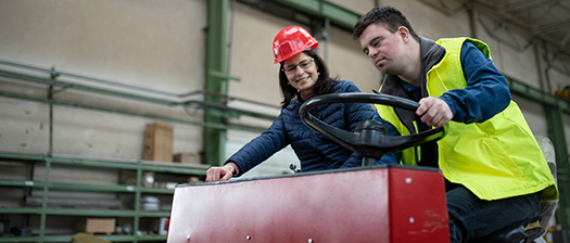 A man with down syndrome wears a hi-vis vest and drives a small vechile around a factory, while a woman wearing a red hard hat sits next to him.