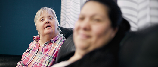 Two women with disability sit on a couch. One looks over the head of the other woman, while the one in the foreground smiles at the camera.