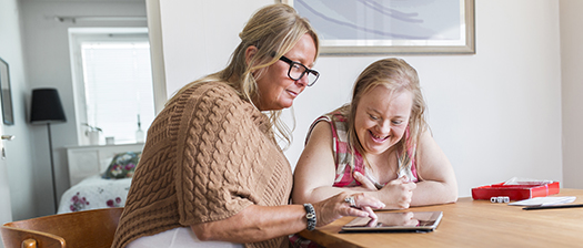 A person with down syndrome and their support person sit at the kitchen table looking at an electronic device