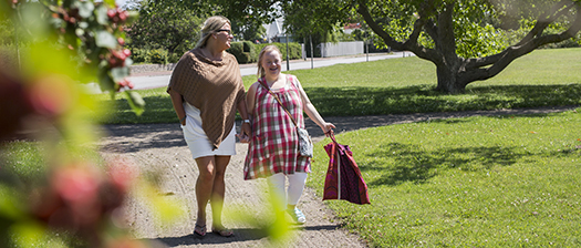 A middle aged woman and a young woman with Down syndrome walk through a sunny park while smiling.