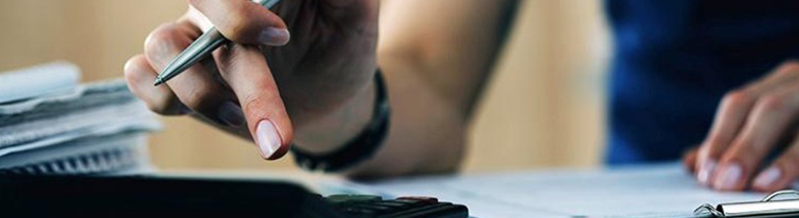 Close up of hand holding a pen and using a calculator on a desk beside paper