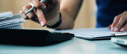 Close up of hand holding a pen and using a calculator on a desk beside paper