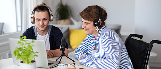A woman and a young man sit in a living room at a computer with a microphone. They both have a set of headphones on.