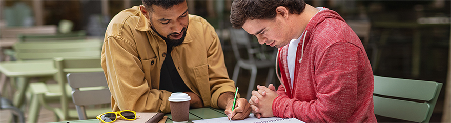 A support worker and a young man with Down syndrome sit outside at a table writing on paper