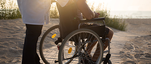 A man assists another man using a wheelchair down a path to a beach. The sun is setting on the beach.