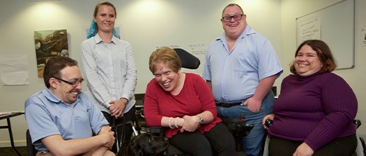 Five people are in a room. Two are standing in the back, and three are using wheelchairs. They are all smiling and laughing. 