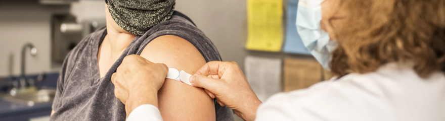 Close up of a nurse applying a Band-Aid to a patient's upper arm after receiving a vaccine injection. They are both wearing surgical masks. 