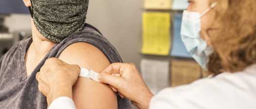 Close up of a nurse applying a Band-Aid to a patient's upper arm after receiving a vaccine injection. They are both wearing surgical masks. 