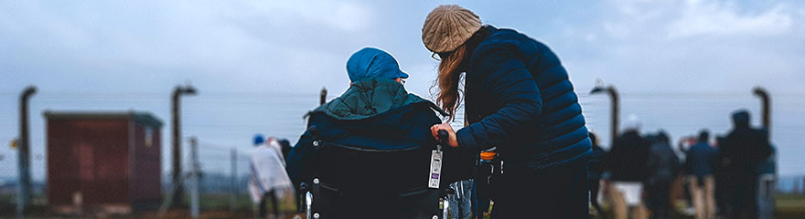 In a field, a woman leans over another woman using a wheelchair. Their backs are to the camera
