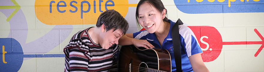 A woman holds a guitar smiling. A person with disability is sitting next to her.