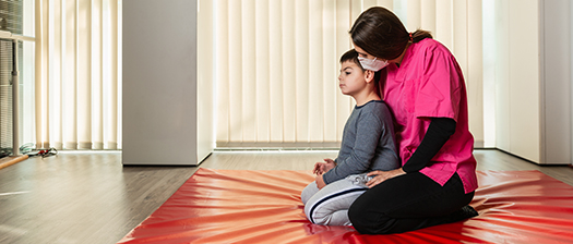 A child sits on a floor mat with a support person sitting behind them
