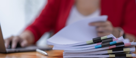 Close up of a stack of document with multicoloured bull clips, a laptop sits in the background
