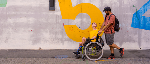 A support person pushing a person with disability past a colourful wall, they are smiling