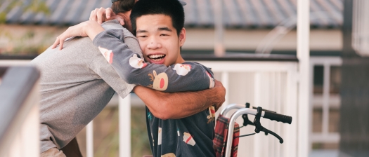 A young person with disability sits in a wheelchair while hugging a support person